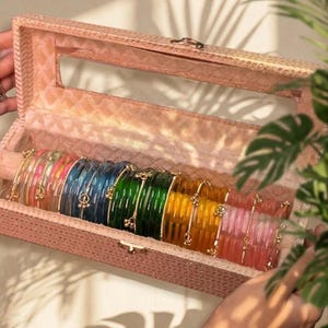 Pode incluir: Uma coleção de pulseiras coloridas numa caixa de joias rosa retangular. As pulseiras são em tons de rosa, azul, verde e amarelo, com detalhes dourados. A caixa está aberta, revelando as pulseiras no interior.