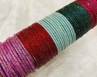 May include: A collection of colorful bangles in shades of purple, red, light blue, green, and pink. Each bangle is adorned with small, sparkling embellishments and gold-colored accents, creating a vibrant and eye-catching display. The bangles are stacked together.