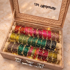 May include: A decorative box with a clear lid, filled with rows of colourful bangles. The bangles are in shades of red, pink, green, and gold. The box is glittery and has gold text on the lid. The bangles are arranged on a cream-coloured fabric.