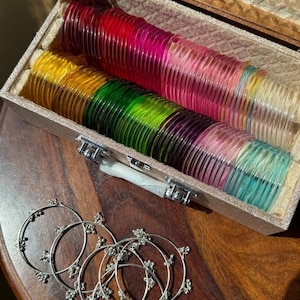 May include: A collection of thin, translucent, multi-colored plastic bangles in shades of red, yellow, green, purple, and blue, stored in a rectangular, textured beige case, alongside several thin silver-toned metal rings with small bead accents.