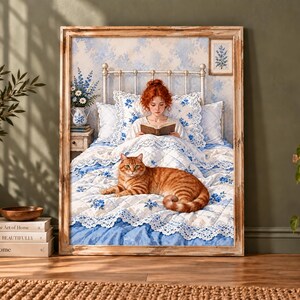 May include: A framed wall art print depicting a person with red hair sitting up in a white bed with blue floral bedding, reading a book. An orange tabby cat lies on the quilt in the foreground. The scene includes a metal bed frame and pillows.