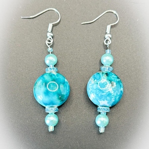 以下が含まれることがあります： 大理石模様の大きなターコイズ色の丸いビーズが特徴の、ぶら下がりタイプのイヤリングです。イヤリングはシルバー色のフック、小さめの水色のビーズ、キラキラ光る装飾が施されています。エレガントなデザインです。