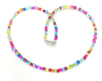 May include: A colorful beaded necklace with a variety of small, square beads in shades of pink, blue, yellow, green, and orange. The necklace has a silver clasp and is displayed on a white background. The beads are strung together to form a necklace.