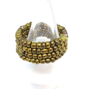 May include: A gold beaded ring with a stack of beads forming a wide band. The ring is displayed on a clear plastic ring holder.