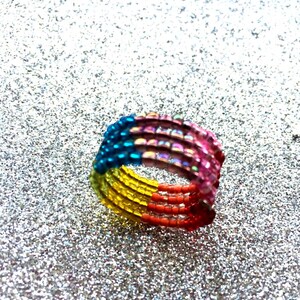 May include: A multi-strand ring constructed from small, cylindrical beads arranged in a gradient pattern. The colours transition from blue to iridescent purple, pink, red, orange, and yellow, forming a continuous circular band of vibrant beads.