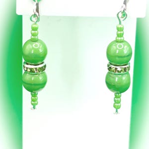May include: A pair of green beaded earrings. Each earring features two large, round green beads separated by a silver band with small green rhinestones. The earrings have a small cluster of green beads at the top and bottom.