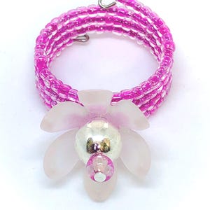 May include: A handmade ring featuring a spiral design of small, magenta beads. The ring is adorned with a light pink flower with a silver center and a smaller pink bead. The ring is a unique piece of jewelry.