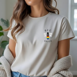 Può includere: T-shirt color crema con una piccola grafica di un'ape con ali arcobaleno e le parole "BEE PROUD" in nero. La maglietta è realizzata in materiale morbido e ha uno scollo tondo.