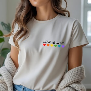 Può includere: T-shirt color crema con la scritta nera "LOVE IS LOVE" e sei cuori arcobaleno sottostanti. La maglietta è a maniche corte e ha uno scollo tondo.