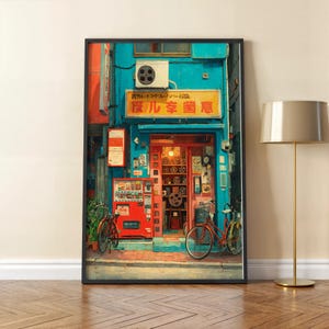 Op de afbeelding: Ingelijste kunstdruk van een Japanse winkel met een turquoise gevel en rode deur. De winkel heeft een automaat en fietsen buiten. Het bord van de winkel heeft Japanse tekst.