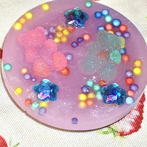 May include: A round, lavender-colored resin piece with embedded glitter, colorful beads, and sparkling accents. Features two glittery teddy bear shapes and three blue, gem-like flower shapes. The piece is decorated with small, colorful spheres.