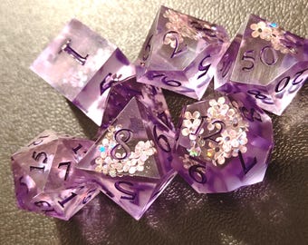 May include: A set of translucent purple polyhedral dice featuring embedded small white flower shapes with iridescent centers. Each die has clear, sharp edges and dark purple ink filled numbers engraved on the faces for readability.