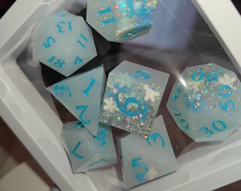 May include: A set of polyhedral dice in a white floating display frame. The dice are pale blue with iridescent glitter and white snowflake inclusions. Each die features bright blue numbers engraved on the surfaces, including d20, d12, d10, d8, d6, and d4 shapes.