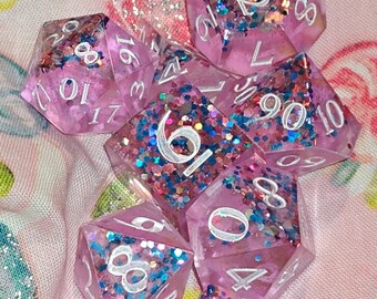 May include: A set of seven polyhedral dice made of translucent pink resin filled with multi-colored glitter and sequins. Each die features white engraved numbers, including a d20, d12, d10, d8, d6, d4, and a percentile die, all arranged in a cluster.