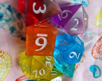 May include: A set of seven translucent, multi-colored polyhedral dice featuring white engraved numbers. The collection includes red, orange, yellow, green, blue, and purple dice of various shapes, including d4, d6, d8, d10, d12, and d20 variants.