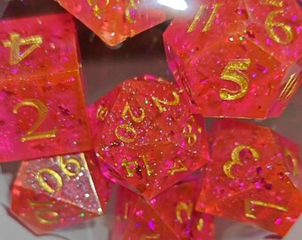 May include: A set of polyhedral dice in a vibrant hot pink color with embedded glitter and gold-colored numbers. The dice feature various geometric shapes including a d20, d12, d10, d8, d6, and d4, all displayed together in a clear container.