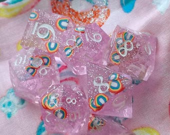 May include: A set of translucent pink polyhedral dice featuring embedded miniature rainbow graphics and silver glitter particles. Each die face displays white numerical digits, with the rainbow designs scattered throughout the resin material.