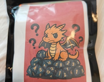 May include: A black plastic resealable pouch containing a label with a red background. The label features an orange cartoon dragon sitting on a pile of dark blue polyhedral dice with gold numbers. The text Mystery Dice Set is printed below.