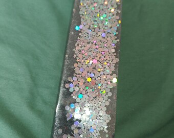 May include: An elongated, rounded rectangular tray made of black resin. The surface is embedded with a dense cluster of multi-sized, holographic hexagonal glitter pieces that reflect various colors, creating a shimmering effect across the center.
