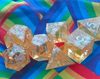 May include: A set of polyhedral dice made from translucent orange resin with embedded white star-shaped glitter and silver-colored numbers. The collection includes various geometric shapes such as a d20, d12, d10, d8, d6, and d4, all featuring sharp edges.