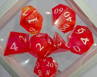 May include: A set of seven polyhedral dice in a translucent red and orange marbled resin with white engraved numbers. The dice set includes a d20, d12, d10, d8, d6, d4, and a percentile die, all displayed within a square white floating frame.