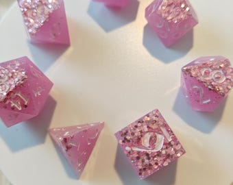 May include: A set of seven polyhedral dice in a translucent pink resin. Each die features a distinct section filled with pink and silver glitter, contrasting with the clear pink resin. The numbers are engraved and filled with a white pigment.