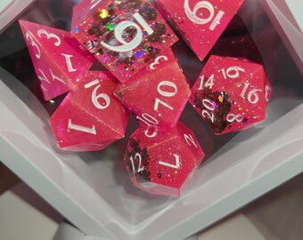 May include: A set of seven polyhedral dice in a white floating display frame. The dice are translucent hot pink with white numbers and embedded dark brown and iridescent glitter inclusions. The set includes various shapes like d20, d12, d10, d8, d6, and d4.