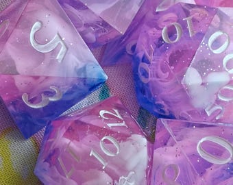 May include: A set of polyhedral dice featuring a translucent resin blend of pink, purple, and blue swirls with embedded silver glitter. Each die has deeply engraved white numbers, including a d20, d12, and d10, showcasing a smooth, polished finish.