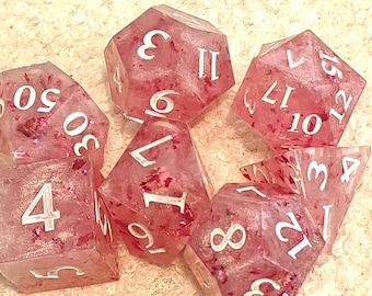 May include: A set of seven polyhedral dice made of translucent pink resin with embedded red glitter flakes. Each die features white engraved numbers. The set includes a d20, d12, d10, d8, d6, d4, and a percentile die, all displaying various numerical values.