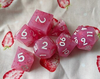 May include: A set of seven polyhedral dice in a translucent pink resin with embedded red glitter flakes and white painted numbers. The collection includes a d20, d12, d10, d8, d6, d4, and a percentile die, all featuring sharp edges and clear numerals.