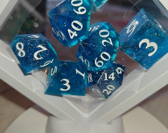May include: A set of seven polyhedral dice in a translucent blue resin with embedded silver glitter, featuring white numbers. The dice are suspended within a white, diamond-shaped floating display frame that is mounted on a circular white base.