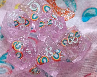 May include: A set of translucent pink polyhedral dice with silver glitter and embedded miniature rainbow motifs. Each die features white engraved numbers. The dice are multi-sided, including d20, d12, d10, d8, d6, and d4 shapes, all crafted from a clear resin material with a sparkling finish.