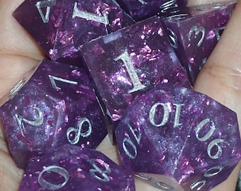 May include: A set of polyhedral dice in a translucent deep purple resin with embedded shimmering purple foil flakes and large, clearly legible silver-colored numbers engraved on each face, including d20, d12, d10, d8, d6, and d4 shapes.