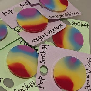 May include: A collection of Pop Socket phone grips with a rainbow gradient design. Each grip is a circular shape with a smooth, glossy finish. The grips are attached to small pink and green cards that read "crafted with love."