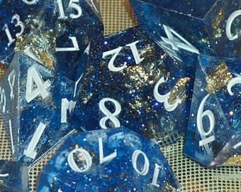 May include: A set of polyhedral dice made from translucent dark blue resin with embedded gold leaf flakes and fine glitter. Each die features sharp edges and clear white painted numbers, including a d20, d12, d10, d8, d6, and d4 for tabletop gaming.