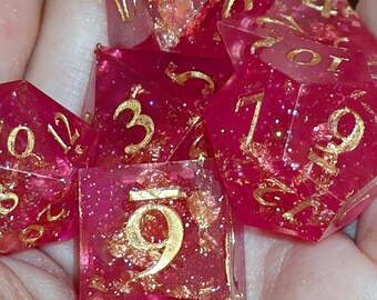 May include: A set of polyhedral dice made from translucent magenta resin with embedded gold-colored metallic flakes and glitter. Each die features engraved numbers painted in a metallic gold finish, including a d20, d12, d10, d8, d6, and d4.