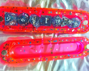 May include: Two elongated, neon orange resin cases with magnetic closures, decorated with multicolored star-shaped glitter. The top case contains a set of seven polyhedral dice in dark blue and purple with silver numbers and embedded glitter.