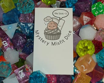 May include: A collection of multi-colored polyhedral dice in various translucent, glittery, and opaque finishes. A white rectangular card is placed in the center with the text Mystery Misfit Dice and an illustration of a cupcake with a die on top.