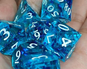 May include: A set of polyhedral dice made of translucent blue resin with embedded dark blue glitter flakes. Each die features sharp edges and clear white engraved numbers. The collection includes various shapes such as a d20, d12, d10, d8, d6, and d4, all displaying a vibrant, sparkling appearance throughout the material.