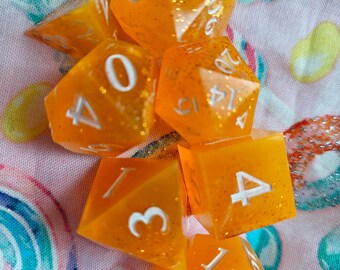 May include: A set of seven translucent orange polyhedral dice with embedded gold glitter and white engraved numbers. The collection includes various geometric shapes such as a d20, d12, d10, d8, d6, and d4, all featuring sharp edges and clear numbering.