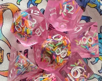 May include: A set of seven translucent pink polyhedral dice featuring embedded multi-colored sprinkles throughout the resin. Each die has large, silver-colored engraved numbers. The collection includes various shapes, such as d20, d12, d10, d8, d6, and d4.