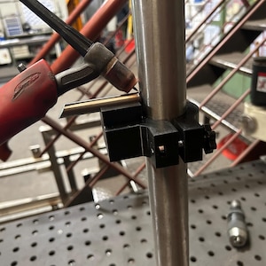 Könnte beinhalten: Nahaufnahme einer Schweißvorrichtung mit einem rot-schwarzen Schweißbrenner in der Nähe einer Metallstange. Eine schwarze Halterung hält ein Metallstück zum Schweißen fest. Der Hintergrund umfasst Metalltreppen und Werkstattausrüstung.