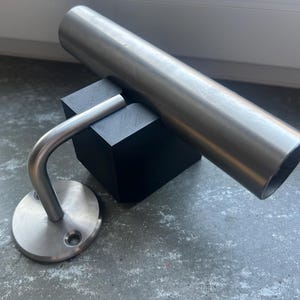 Könnte beinhalten: Eine Edelstahl-Handlaufstütze mit gebogenem Arm und kreisförmiger Basis. Ein zylindrischer Metallhandlauf liegt auf einem schwarzen quadratischen Block. Handlauf und Stütze sind aus gebürstetem Metall.