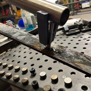 May include: A metal tube held by a black clamp, positioned over a metal plate with a grid of holes and metal fixtures. The image suggests a workshop or industrial setting. The tube is made of metal.