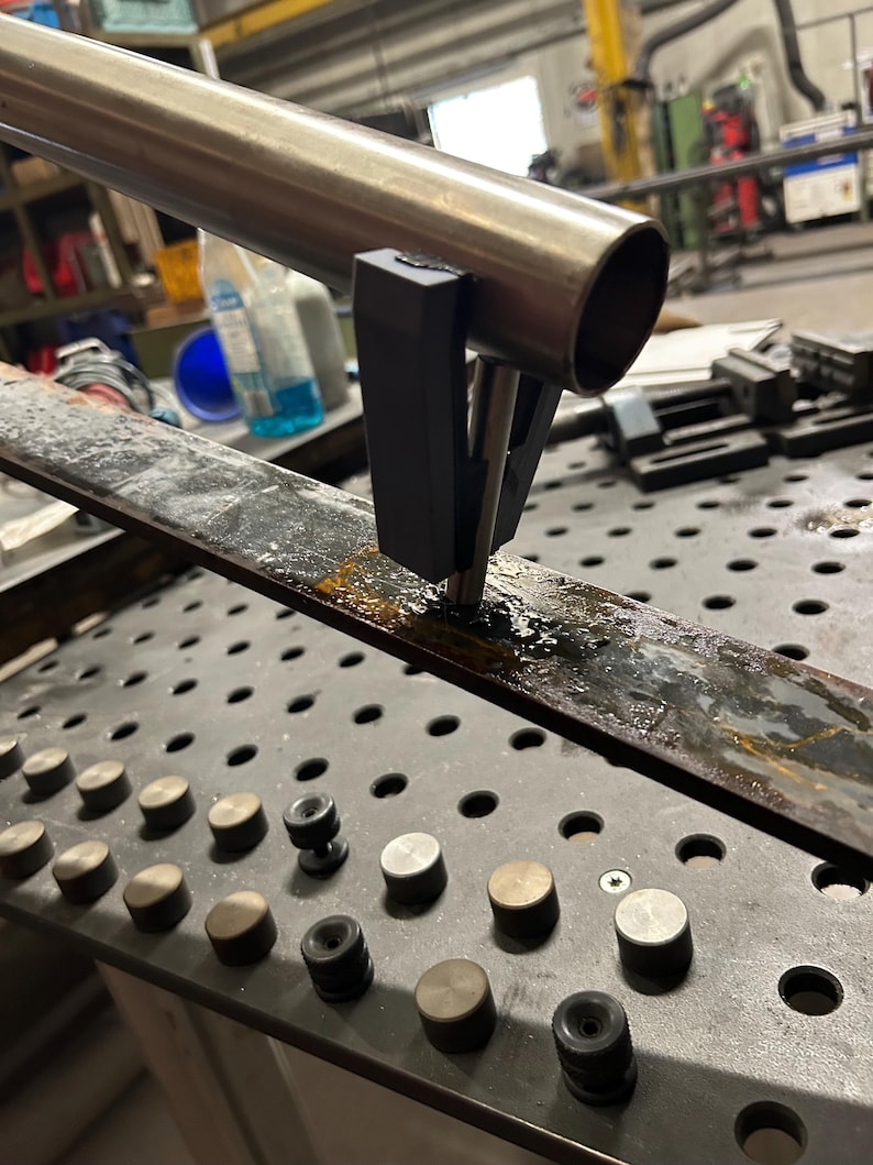 May include: A metal tube held by a black clamp, positioned over a metal plate with a grid of holes and metal fixtures. The image suggests a workshop or industrial setting. The tube is made of metal.