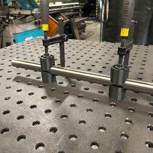 Könnte beinhalten: Eine Edelstahl-Metallstange wird von zwei schwarzen Klemmen auf einer perforierten Metallarbeitsfläche gehalten. Die Klemmen haben gelbe Akzente. Werkstattumgebung.