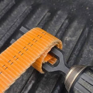 Könnte beinhalten: Ein schwarzes Metallwerkzeug zum Aufwickeln von Gurten mit einem Y-förmigen Schlitzende, das in eine Schlaufe aus leuchtend orangefarbenem, strapazierfähigem Nylongewebe eingeführt ist. Das Werkzeug hat einen strukturierten Griff.