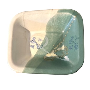 Puede incluir: Un plato cuadrado de cerámica con un diseño bicolor. Un lado es blanquecino, el otro es verde-azulado apagado. El plato presenta un sutil patrón floral en azul y detalles dorados, adecuado para servir o exhibir.