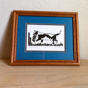 May include: A framed black and white print of a dog carrying an object in its mouth. The dog has black spots and is set against a white background with grass. The print is matted in blue and framed in wood.