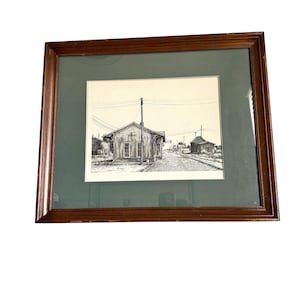 May include: A framed black and white pen and ink drawing of a small town scene. The artwork depicts a building with the word "STUART" on it, power lines, and other buildings. The frame is a dark brown color, and the matting is green.